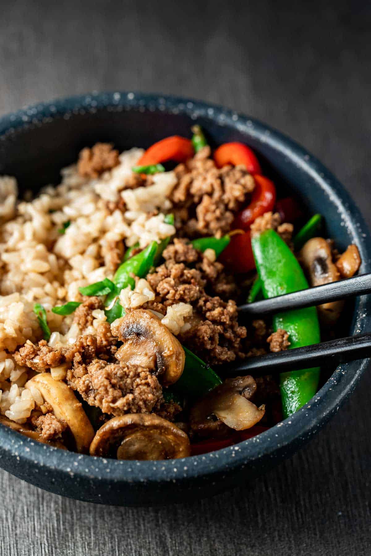 Ground beef stir fry in a bowl with rice and chop sticks.
