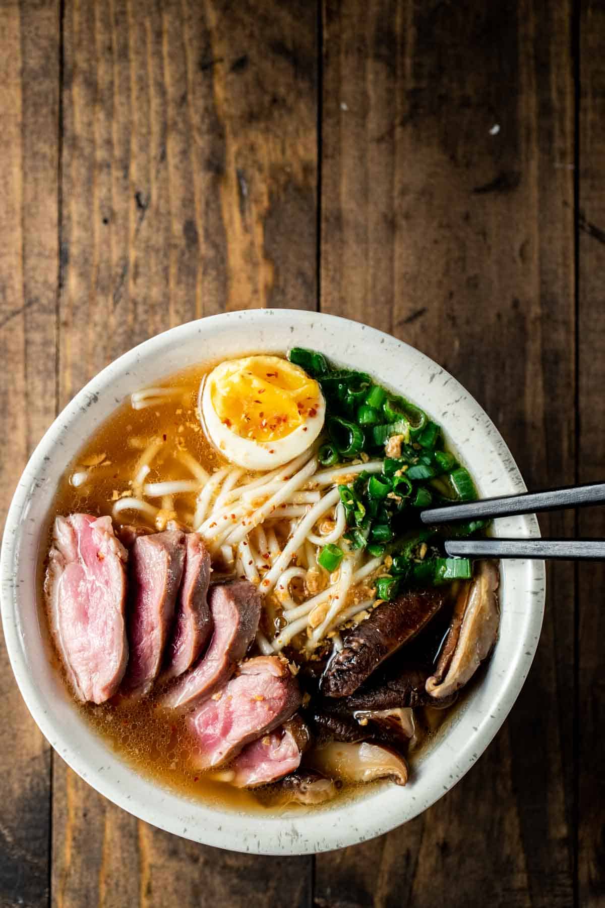 up close view of chop sticks in a white bowl with duck, ramen, scallions, and broth