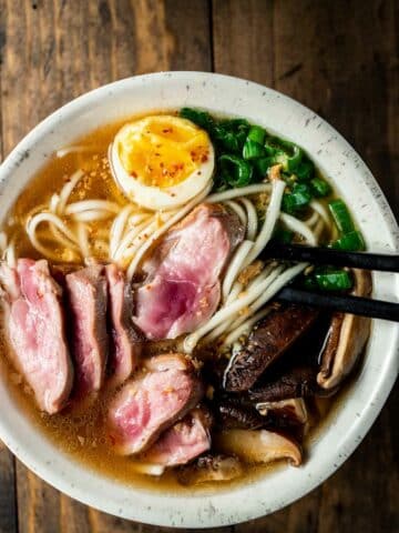 view of duck ramen in a bowl with eggs, duck, noodles, scallions, with black chop sticks