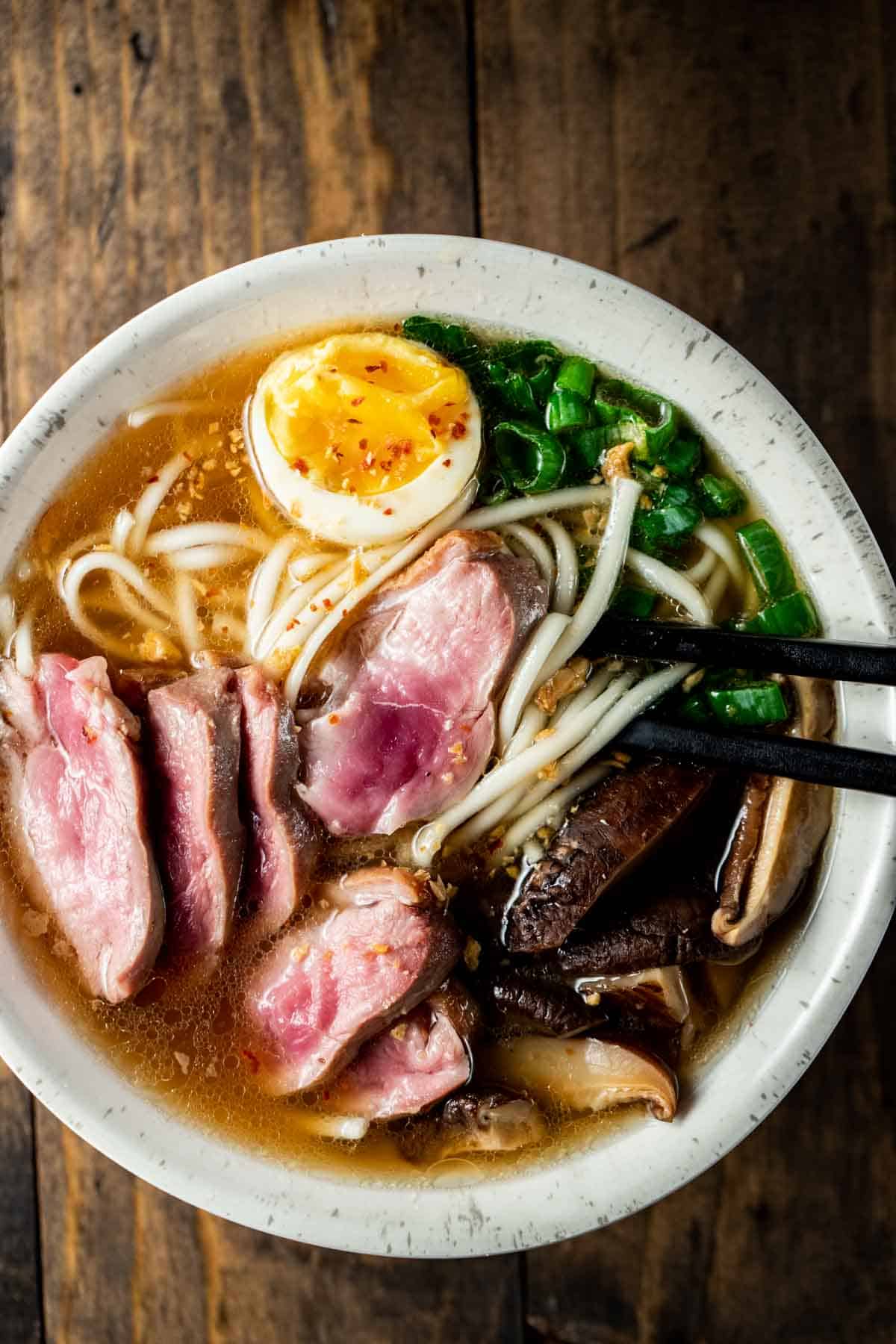 view of duck ramen in a bowl with eggs, duck, noodles, scallions, with black chop sticks