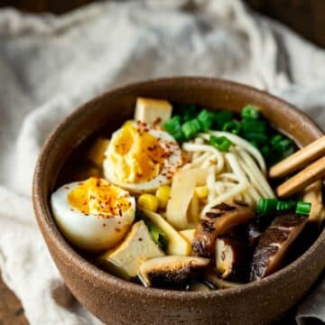 cooked ramen, eggs, vegetables, and meat in a brown bowl with chopsticks on a white towel