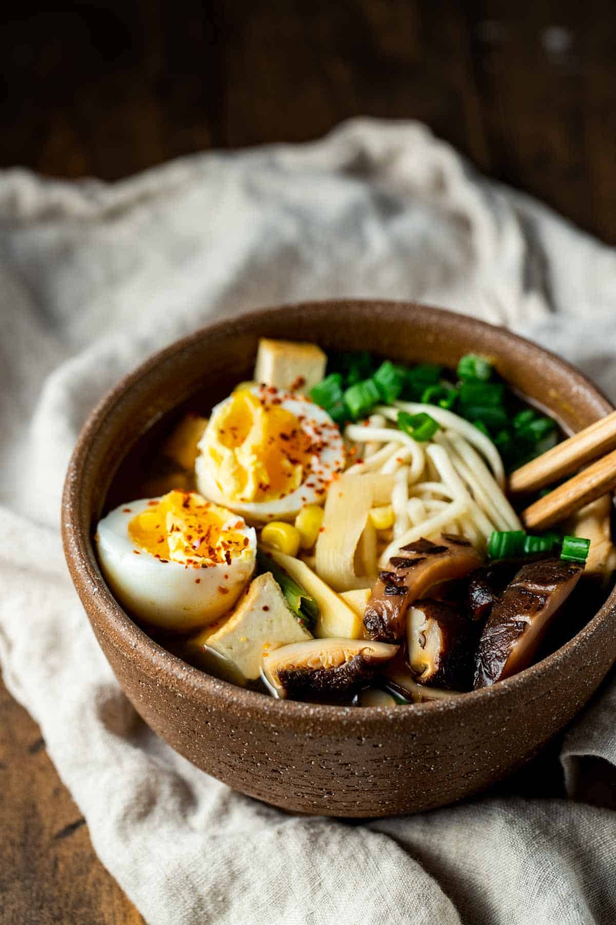 cooked ramen, eggs, vegetables, and meat in a brown bowl with chopsticks on a white towel