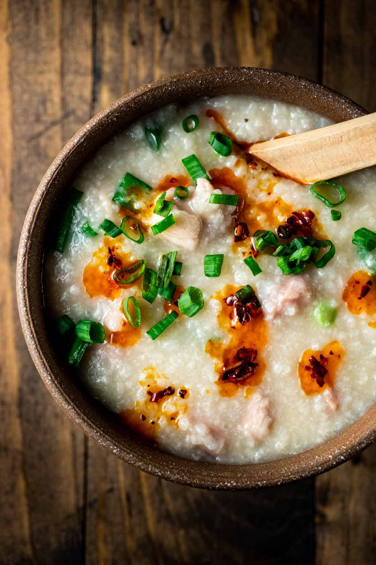a brown bowl of congee with a wooden spoon in it with green onions and chili oil on top
