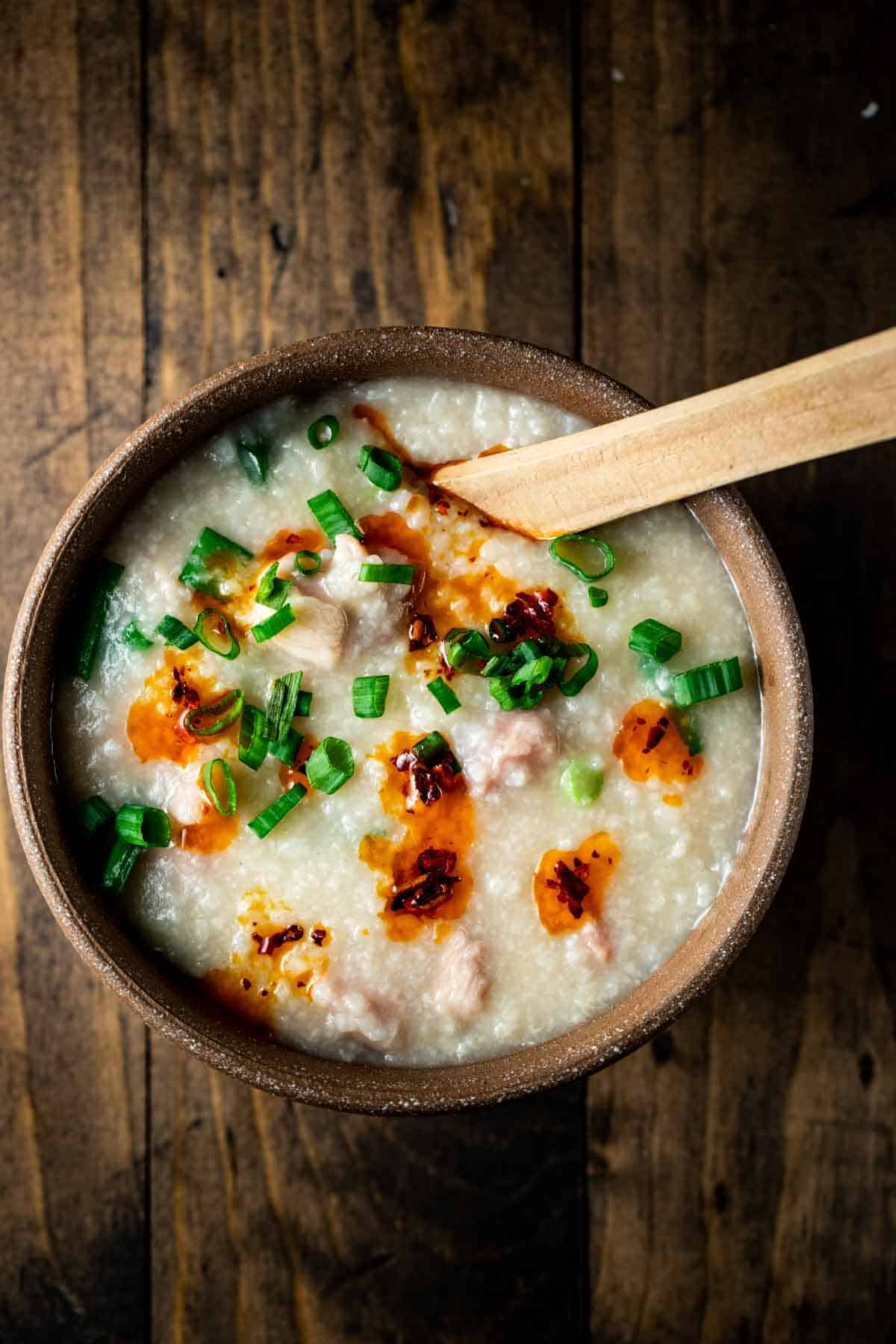 a bowl of congee with chili oil and green onions on top on a wooden table