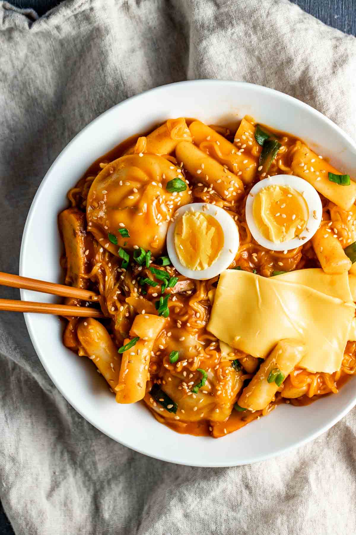 korean rice cakes, american cheese slices, noodles, eggs and chives in a white bowl with chopsticks