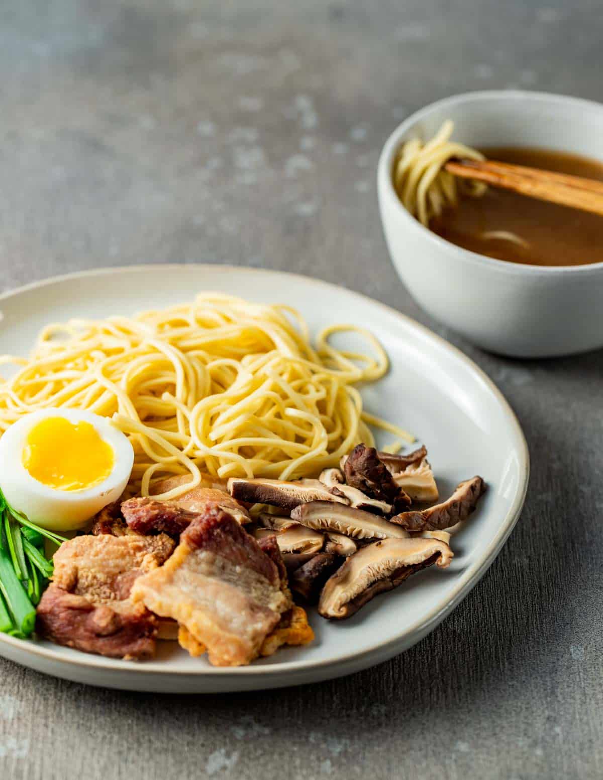 a plate of pork belly and noodles with a bowl of broth and chopsticks in the background