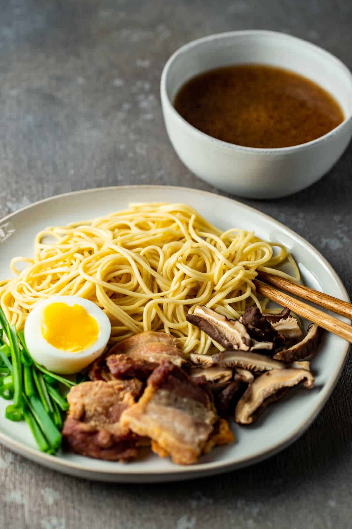 chopsticks on a plate with noodles, pork belly, egg and green onions awith a bowl of broth in the background