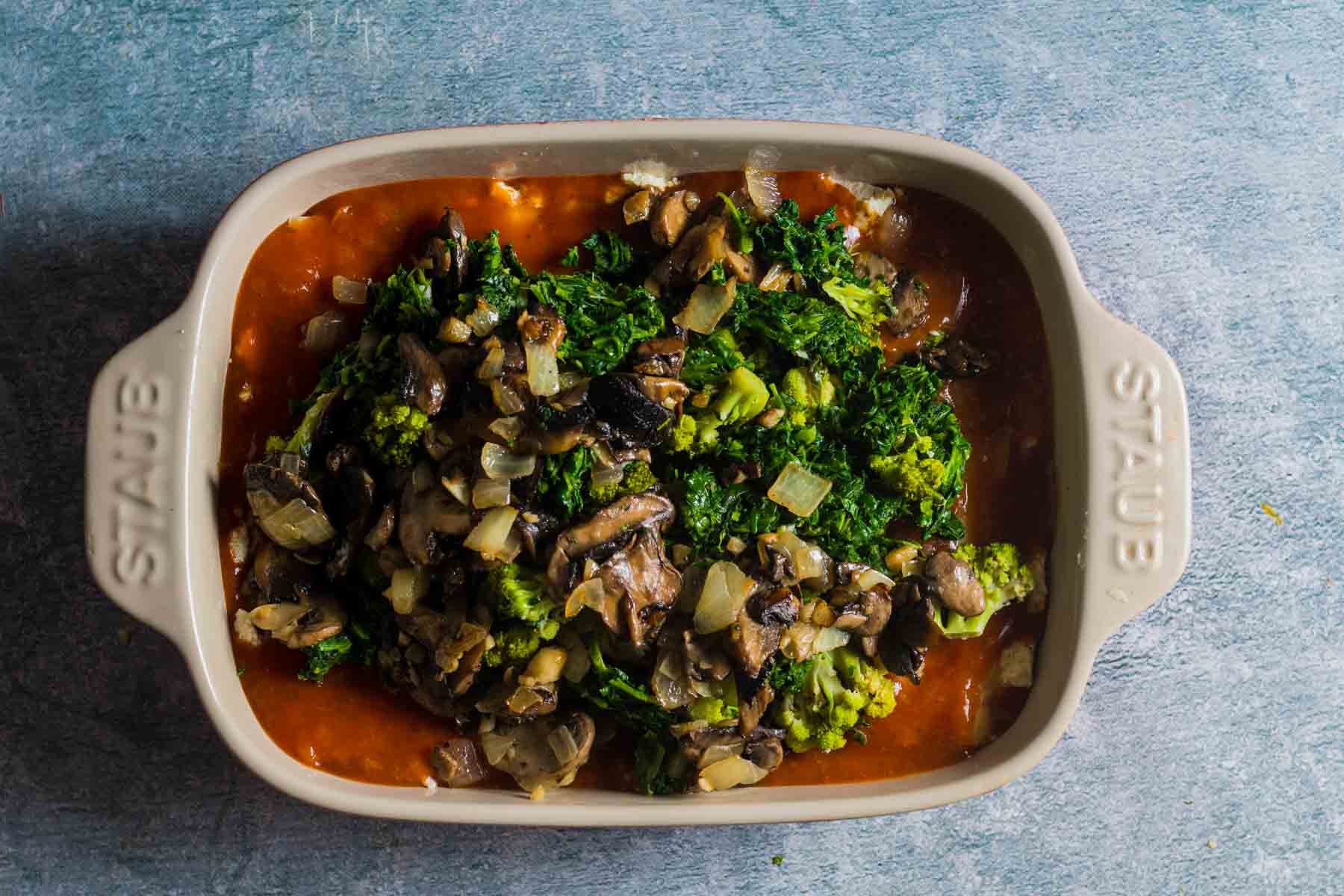 green vegetables and mushrooms on top of red sauce in a baking dish