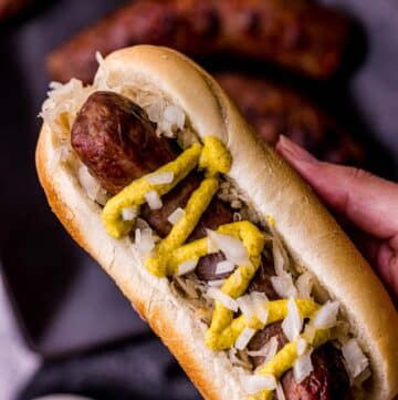 a hand holding a sausage on a bun with mustard and onions