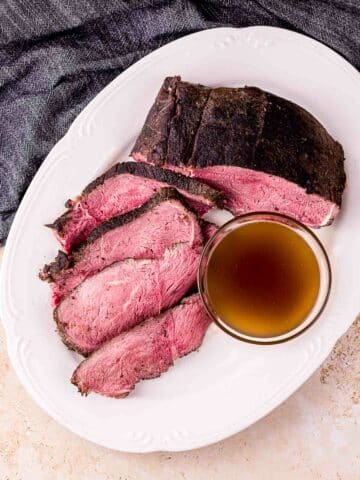 a sliced beef roast on a plate with a bowl of jus