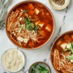 2 bowls of red colored soup with green onions and sesame seeds on the side