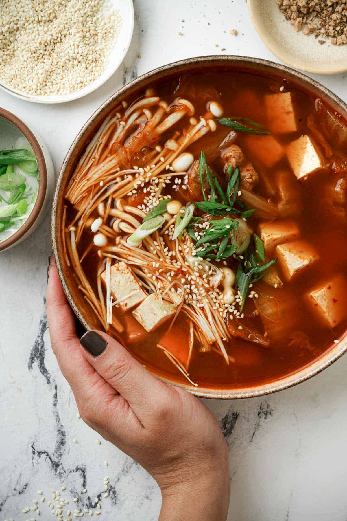 a hand holding a bowl of red colored tofu soup with mushrooms and onions