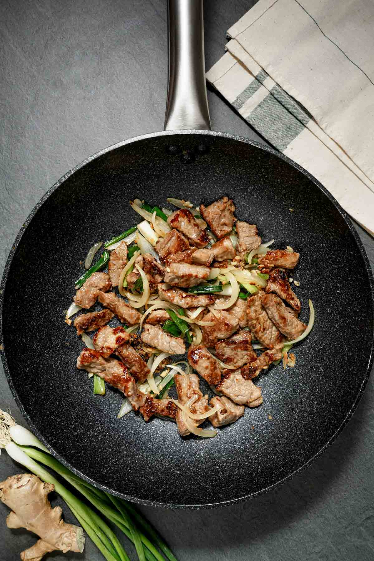 slices of meat, onions and green onions cooking in a skillet