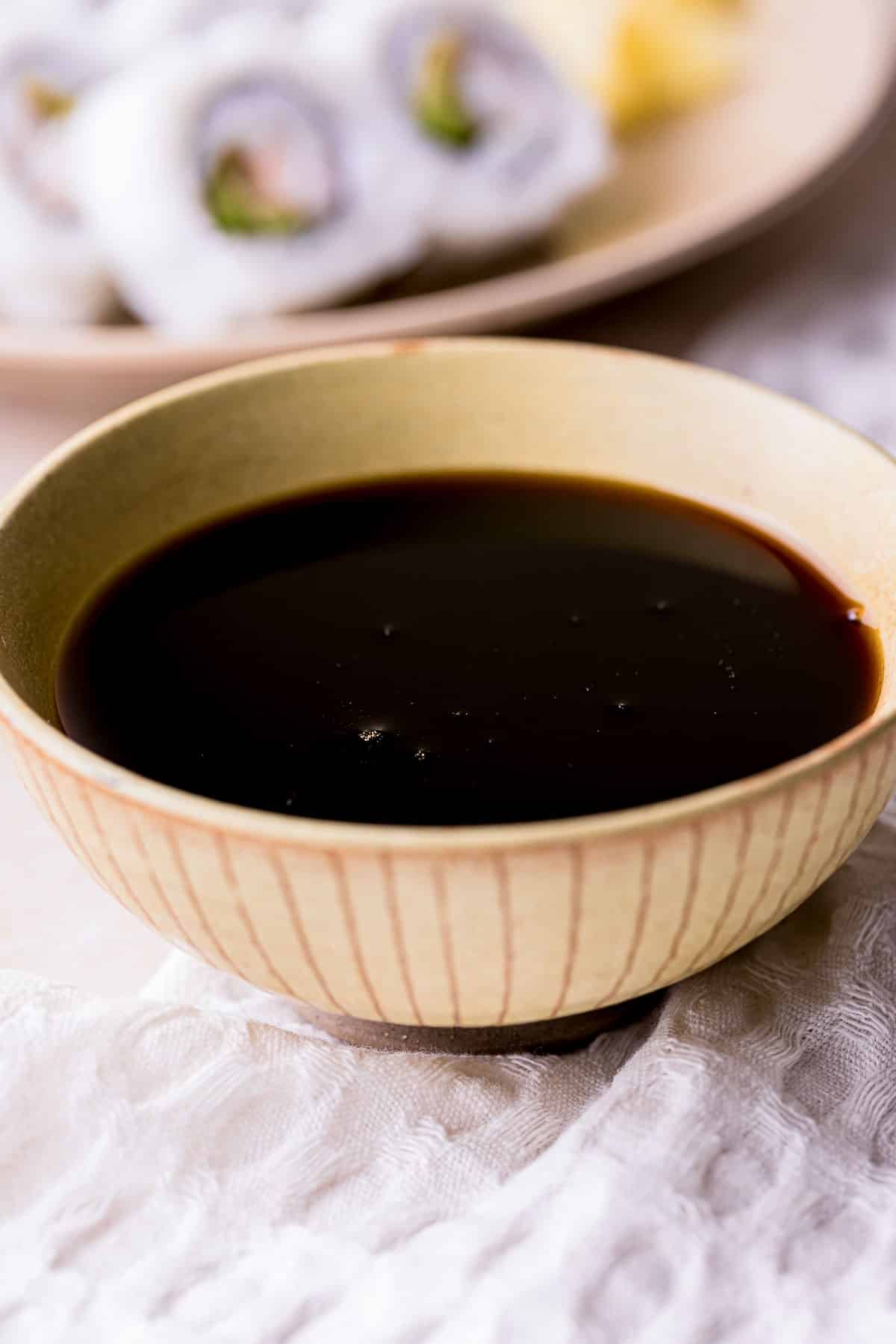 a bowl of dark brown sauce with sushi rolls in the background