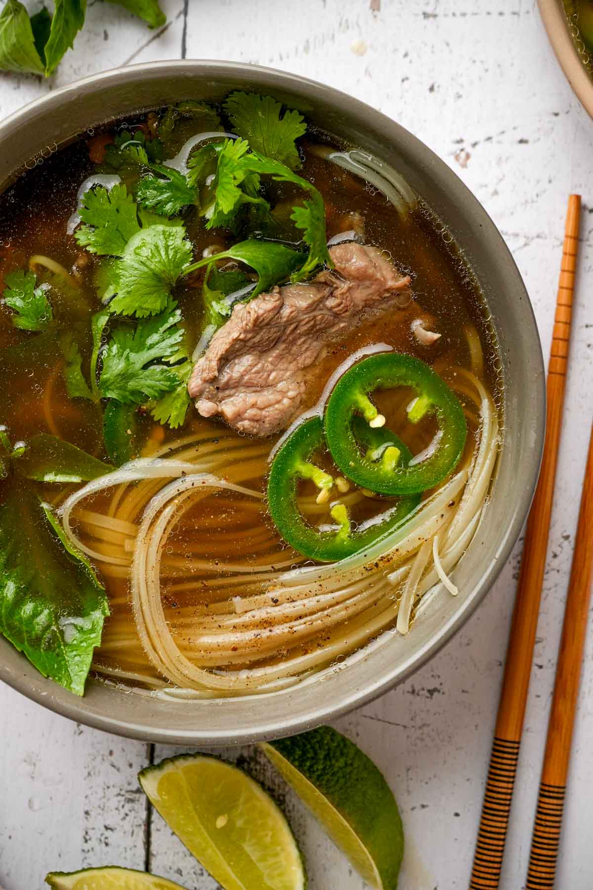 a bowl of pho tai garnished with herbs, jalapenos and pieces of beef