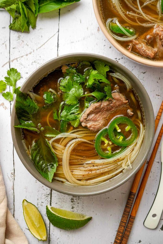 rare beef noodles soup in a bowl with green herbs and slices of pepper