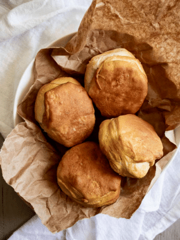 The biscuits have a rounded, slightly uneven shape with visible flaky layers on the sides. Their tops are a deep, toasted gold, suggesting they are fresh from the oven or an air fryer. The bowl sits atop a white, textured linen cloth, creating a cozy, rustic aesthetic.