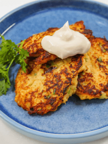 A stack of three crispy, golden-brown vegetable fritters on a blue plate, topped with a smooth dollop of sour cream and garnished with a sprig of fresh parsley.