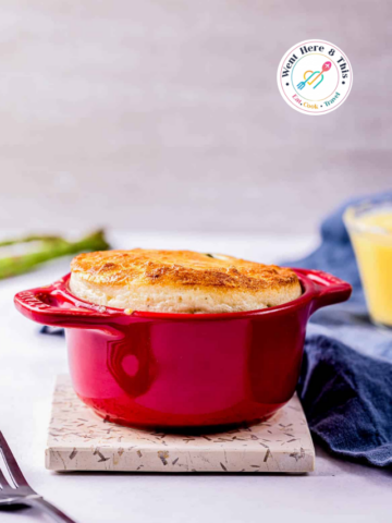 A close-up shot of a creamy asparagus soufflé in a red Staub cocotte, with a black spoon lifting a bite to reveal tender green asparagus pieces and a fluffy, cheesy interior.