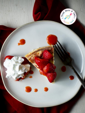 A top-down view of a slice of Creamy Strawberry Shortcake Cheesecake on a white plate, topped with fresh strawberries and strawberry sauce, served with a side of whipped cream and a fork.