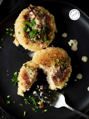 Two crispy golden-brown pan-fried risotto cakes topped with savory mushroom sauce and fresh parsley on a matte black plate with a silver fork.