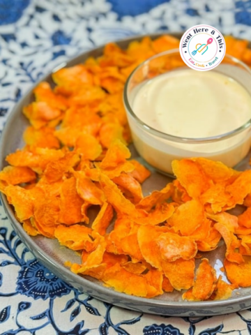 Homemade dehydrated sweet potato chips arranged on a grey plate around a glass bowl of creamy orange dipping sauce, set on a blue patterned tablecloth.