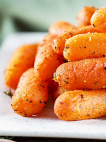 A close-up, tempting stack of roasted baby carrots on a white platter, glistening with a savory glaze and speckled with fresh herbs and cracked black pepper.