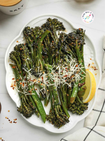 A plate of tender charred broccolini topped with grated parmesan cheese and red chili flakes, served with a fresh lemon wedge on a white scalloped platter.