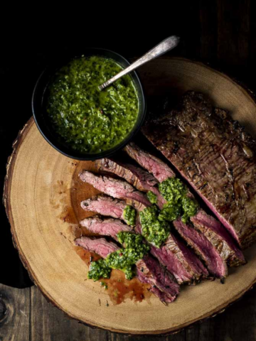 Sliced medium-rare flank steak topped with vibrant green chimichurri sauce, served on a rustic wood slab next to a small bowl of extra sauce.
