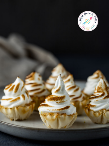 Seven mini lemon meringue tarts are arranged on a simple grey ceramic plate against a dark, moody background. Each tart features a golden-brown, flaky phyllo pastry shell filled with lemon curd and topped with a high swirl of toasted white meringue. The torched edges of the meringue create a beautiful brown-and-white contrast, emphasizing the light and airy texture.