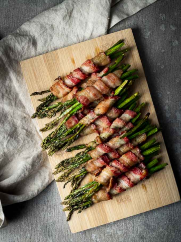 Crispy bacon-wrapped asparagus spears arranged in rows on a light wood cutting board, served next to a linen napkin.