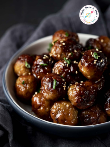 A close-up of a bowl filled with glossy, glazed air fryer meatballs coated in a sweet and spicy sticky sauce, garnished with toasted sesame seeds and minced green chives.