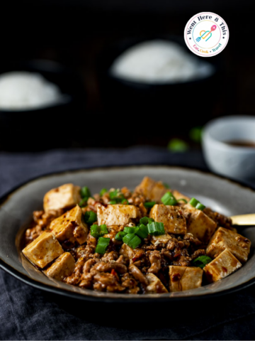 A bowl of authentic spicy Sichuan Mapo Tofu with soft silken tofu cubes, savory minced pork, and spicy chili oil, garnished with fresh green onions, served with side bowls of white rice on a dark table.