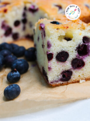 A close-up shot of a thick slice of fluffy blueberry breakfast cake on parchment paper, filled with juicy, ripe blueberries and surrounded by fresh loose berries.