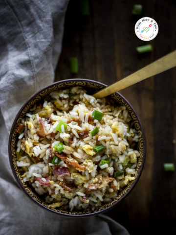 A ceramic bowl of savory breakfast fried rice with scrambled eggs, crispy bacon bits, and fresh green onion garnish on a rustic wooden table.