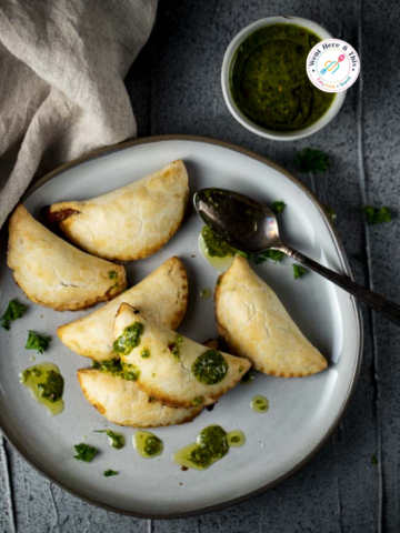 A plate of five golden-brown baked vegetarian empanadas drizzled with vibrant green chimichurri sauce. A small bowl of extra sauce and a spoon are nearby on a textured grey background.