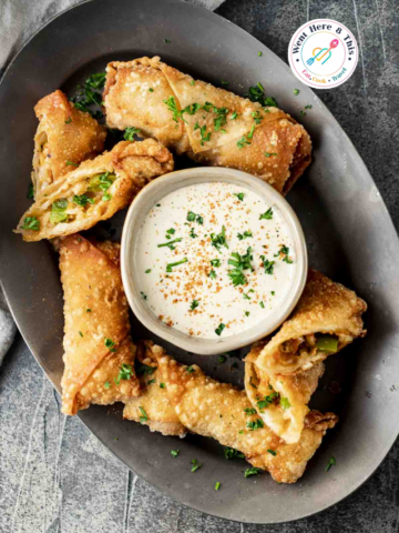 Crispy golden-brown Cajun crawfish egg rolls cut on a bias, arranged on a dark metal platter around a white dipping bowl filled with seasoned cream sauce and fresh parsley.
