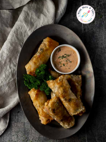 Top-down view of a dark metal platter holding golden, crispy Southwest Egg Rolls served with a side of creamy chipotle dipping sauce and fresh green parsley garnish.