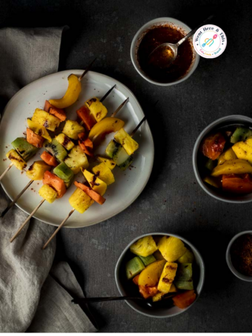 Top-down view of grilled fruit kabobs on skewers featuring pineapple, peach, and kiwi with visible char marks. Served on a white plate next to small bowls of chili lime seasoning and dipping sauce on a dark moody background.