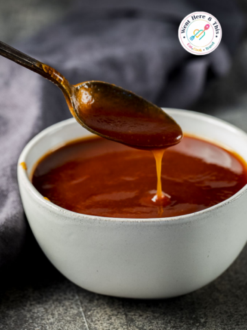 Thick, glossy reddish-brown crispy homemade barbecue sauce dripping from a vintage metal spoon into a white ceramic bowl. High-contrast food photography with a dark, moody background.