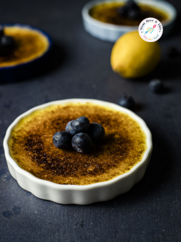 A white ramekin filled with silky lemon crème brûlée, featuring a perfectly torched, caramelized sugar crust topped with fresh blueberries. A whole lemon and additional ramekins are blurred in the dark, moody background.