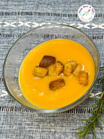 A glass bowl of creamy, golden-yellow vegan potato soup topped with crispy seasoned croutons, served on a blue textured placemat with a sprig of fresh rosemary.