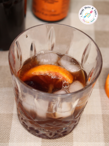 A non-alcoholic Old Fashioned mocktail served in a crystal rocks glass over clear ice cubes, garnished with a fresh orange slice, sitting on a neutral checkered napkin.