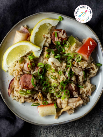 Top-down view of a seafood rice plate garnished with green onions, sausage slices, imitation crab, and fresh lemon wedges.