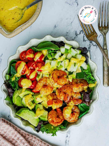 Spicy blackened shrimp salad topped with a vibrant yellow mango dressing, served with diced avocados, fresh cherry tomatoes, and crisp green apples in a fluted ceramic bowl.