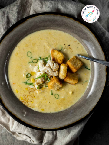 A top-down view of a rustic bowl filled with creamy yellow spicy crab and corn bisque, garnished with fresh lump crab meat, golden croutons, and sliced green onions on a linen napkin.