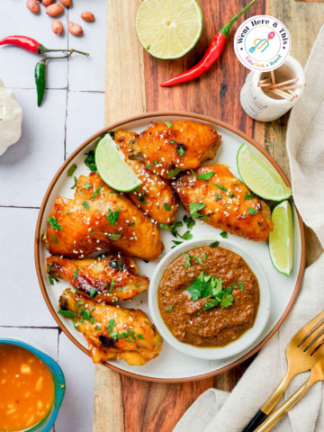 Close-up of sticky Thai chicken wings with lime wedges and a bowl of peanut satay dipping sauce.