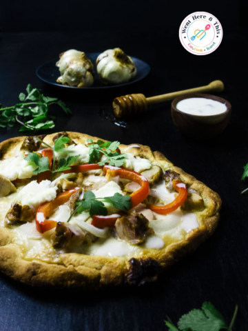 A freshly baked Tandoori Chicken Pizza served on a dark background. The pizza crust is topped with chunks of spiced tandoori chicken, melted white cheese, sliced red bell peppers, onions, and fresh cilantro leaves. In the background, two roasted garlic bulbs sit on a black plate next to a small wooden bowl of white sauce and a honey dipper. A circular "Went Here 8 This" logo is visible in the top right corner.