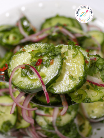 A close-up shot of a silver spoon lifting a fresh serving of Thai Cucumber Salad. The salad is made with thinly sliced crisp green cucumbers coated in a shiny, clear dressing. It is tossed with bright red onion slivers, finely chopped fresh cilantro, and small flecks of red chili peppers. A blurred white bowl filled with more cucumber salad sits in the background.