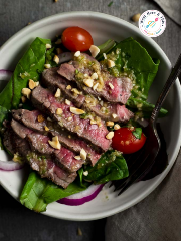 Slices of medium-rare grilled sirloin steak on a bed of fresh spring mix green salad, garnished with chopped peanuts, red onion slices, and cherry tomatoes, drizzled with a zesty garlic lime Thai dressing in a white bowl.